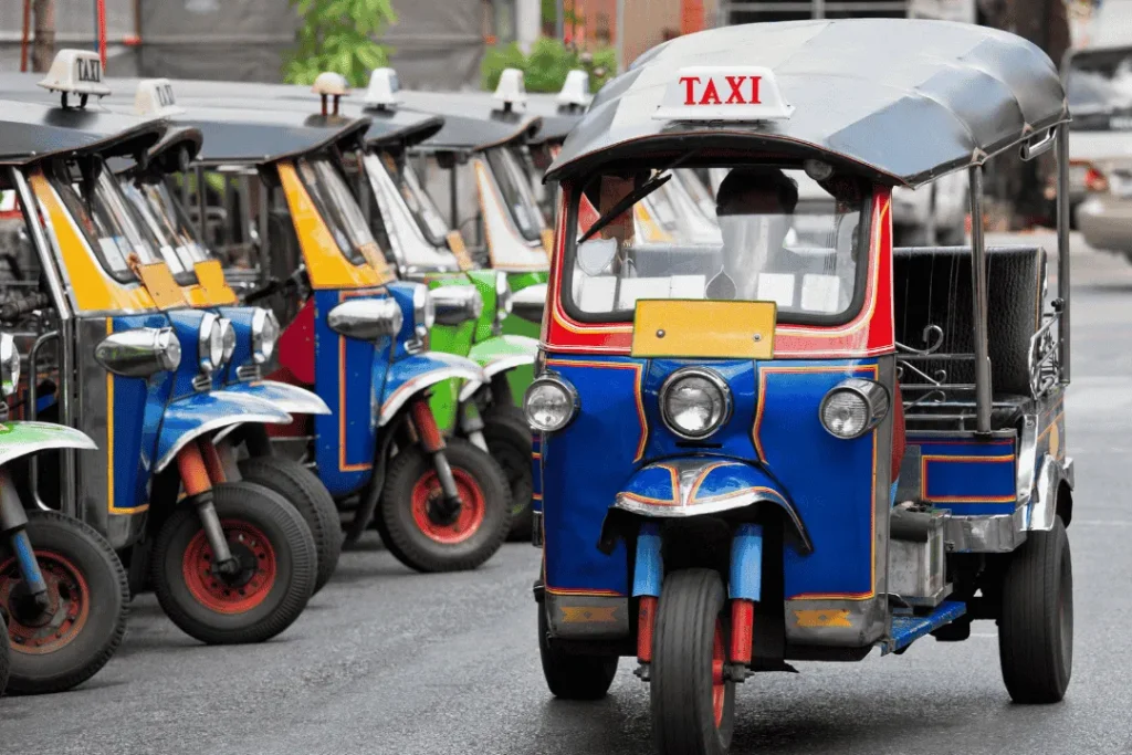 Tuk tuk warna-warni melintas di jalanan Bangkok siang hari dengan suasana kota yang ramai.